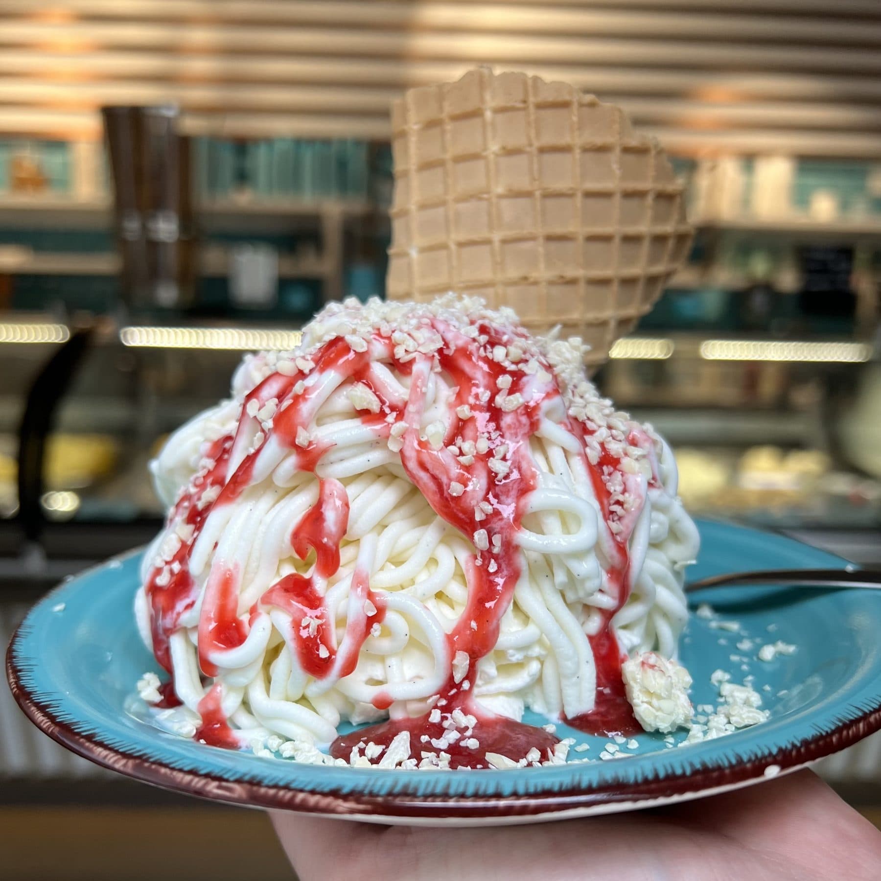 Spaghettieis aus Bourbon-Vanilleeis mit Erdbeersoße, weißen Schokoraspeln und Waffel auf türkisem Teller
