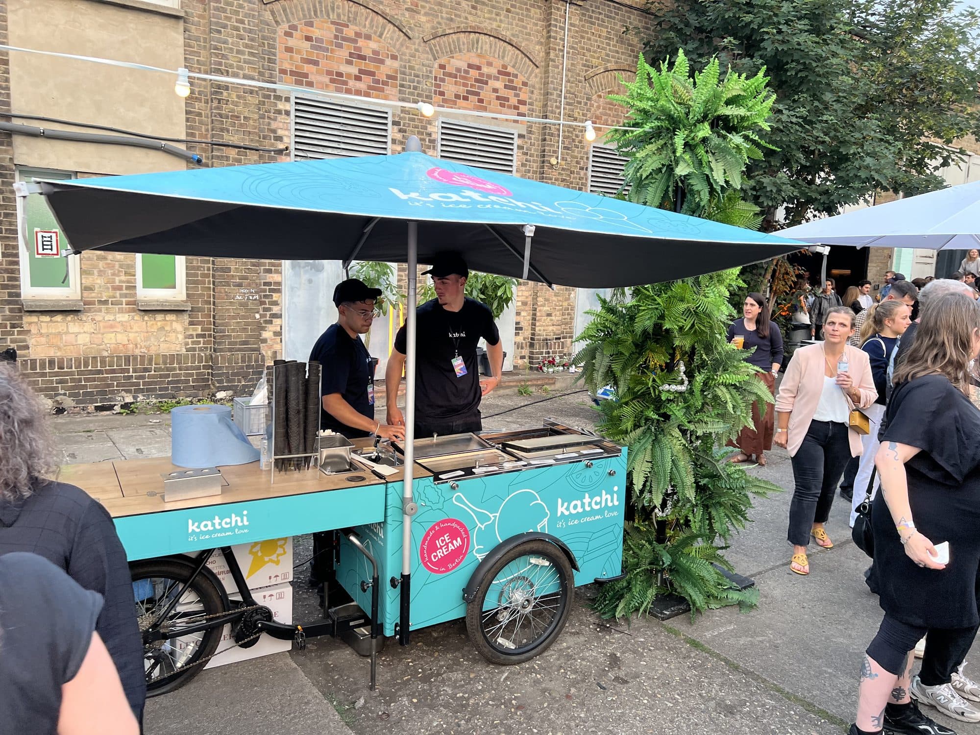 Zwei Katchi-Mitarbeiter in Katchi-Caps und -Shirts am Ice Cream Bike mit Sonnenschirm beim Outdoor-Event