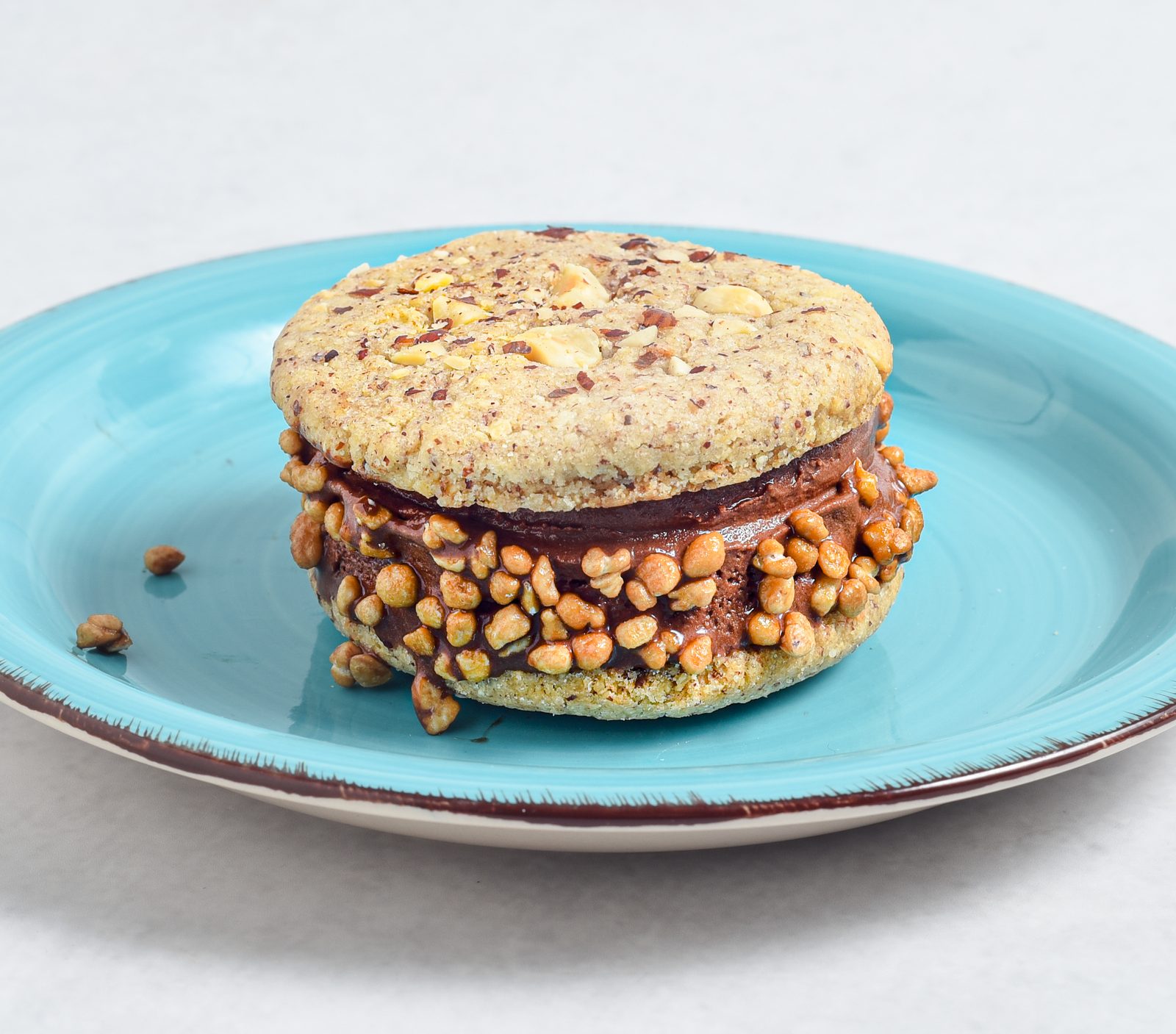 Ice Cream Sandwich mit Schokoladeneis zwischen zwei Nuss-Cookies, der Rand in Krokant gewälzt, auf türkisem Teller