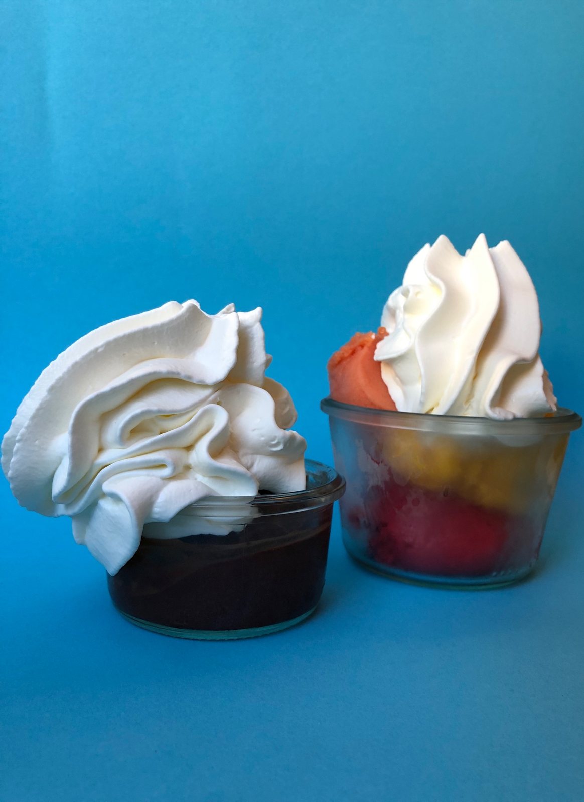 Zwei Dessertgläser mit Schokoladeneis, Fruchtsorbet und Sahnehaube vor blauem Hintergrund