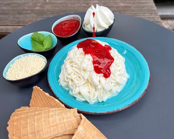 Erdbeersoße, wie sie auf das Spaghettieis drippt und seitlich am Bourbon-Vanilleeis runterläuft. Um den Teller herum stehen die Zutaten für das Spaghettieis: Weiße Schokostreusel, Minzblätter, Erdbeersoße und Schlagsahne.